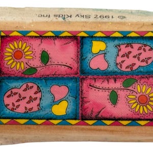 Op de afbeelding: Een houten stempel met een kleurrijk ontwerp met bloemen en harten. Het ontwerp is verdeeld in vier vierkanten met roze en blauwe achtergronden, omlijnd in geel en blauw. De stempel heeft de tekst "© 1997 Sky Kids Inc."