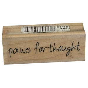 May include: A rectangular wooden rubber stamp featuring the black text "paws for thought" printed on the front side. A white adhesive label with a black barcode and the alphanumeric code 006-11435 56164-8 is attached to the top edge of the wood block.