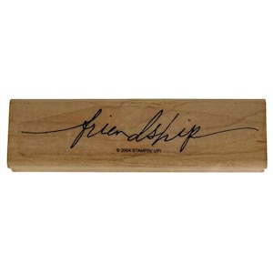 Puede incluir: Un sello de goma rectangular de madera con la palabra friendship escrita en letra cursiva negra en el centro. El texto 2004 STAMPIN' UP! está impreso en letras negras pequeñas justo debajo de la palabra friendship sobre la madera marrón clara.