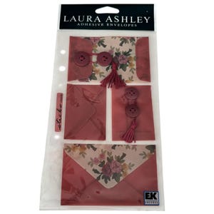 May include: A package of Laura Ashley adhesive envelopes featuring floral patterns and solid dusty rose colors. The set includes envelopes decorated with small buttons and matching tassels, all contained within a clear plastic display sleeve.