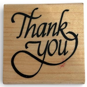 May include: A square wooden rubber stamp featuring the words Thank you in a black, elegant, flowing cursive script. The natural light-colored wood grain is visible across the surface of the stamp block, which has a smooth, flat finish.
