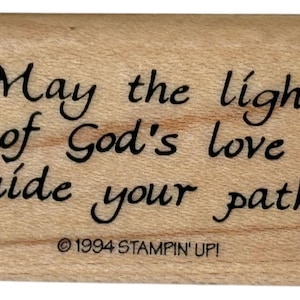 Puede incluir: Un sello de madera rectangular con la inscripción "May the light of God's love guide your path" en cursiva. El sello también incluye la información de copyright "©1994 STAMPIN' UP!"