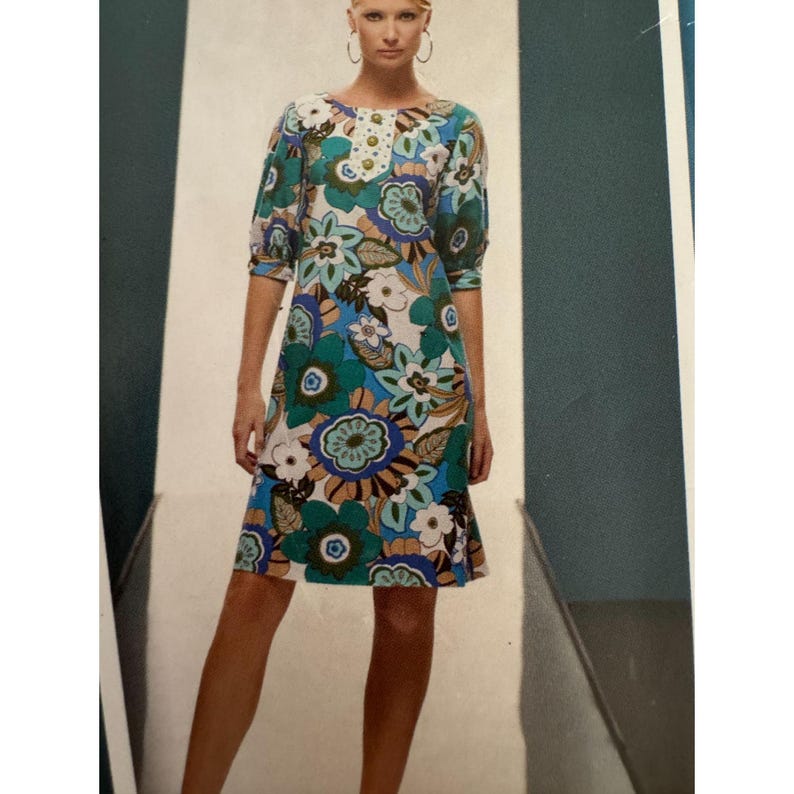 May include: A knee-length dress featuring a vibrant floral pattern in shades of teal, blue, brown, and white. The garment has three-quarter length sleeves with gathered cuffs and a central placket at the neckline adorned with a row of round buttons.