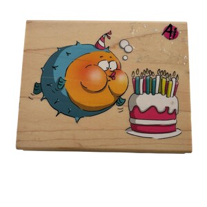 Könnte beinhalten: Ein Holzstempel mit einem Cartoon-Kugelfisch, der Kerzen auf einer Geburtstagstorte auspustet. Der Kugelfisch ist orange und blau und trägt eine Partyhut. Die Torte ist rosa und weiß mit brennenden Kerzen.