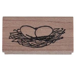 Peut inclure: Un tampon en caoutchouc rectangulaire en bois avec un dessin au trait à l'encre noire représentant deux œufs ovales reposant dans un nid d'oiseau. Le tampon présente une finition en bois clair et une surface supérieure plate.