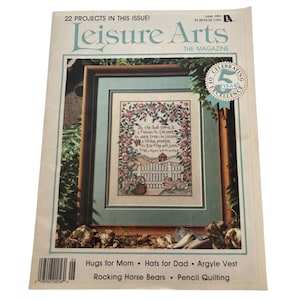 Puede incluir: Una portada de revista Leisure Arts vintage de junio de 1991. La portada presenta un diseño de punto de cruz enmarcado con un borde floral y una cita. El título de la revista está en verde azulado, con el texto "¡22 PROYECTOS EN ESTE NÚMERO!" y "LA REVISTA".