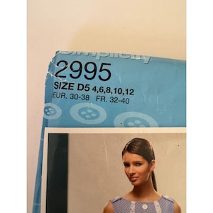 May include: A light blue Simplicity sewing pattern envelope labeled 2995, featuring size D5 with ranges 4, 6, 8, 10, 12, EUR 30-38, and FR 32-40. The packaging displays a pattern of white buttons and a photograph of a person wearing a polka dot garment.