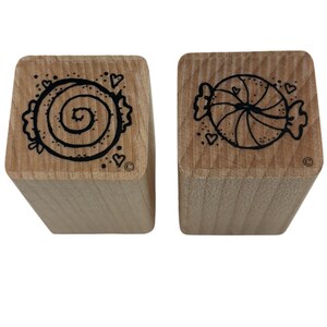 May include: Two wooden stamps with black candy designs. One stamp features a spiral candy with hearts, and the other shows a wrapped candy with hearts. The stamps are on a white background.