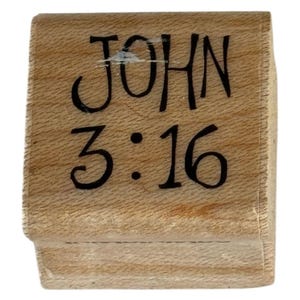 Puede incluir: Un sello de madera con el texto "JOHN 3:16" en negro, con letras estilizadas. El sello tiene forma rectangular y un acabado de veta de madera natural. El texto está centrado en la cara del sello.