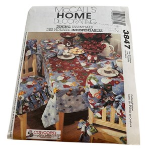 May include: McCall's Home Decorating pattern for dining essentials. The pattern includes a tablecloth, chair covers, and bows. The fabric has a blue background with snowmen and snowflakes. The pattern number is 3847.