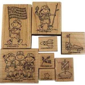 Puede incluir: Un conjunto de sellos de madera con personajes de dibujos animados de exploradores e imágenes patrióticas. Los diseños incluyen un explorador con una bandera, un explorador con una mochila, un cohete, un coche con una bandera y más. Los sellos están marcados con "2002 STAMPIN' UP!"