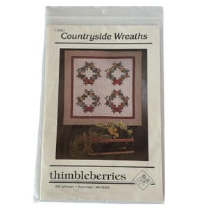 May include: A sealed plastic bag containing a sewing pattern titled "Countryside Wreaths" by thimbleberries. The pattern features a quilt design with four wreath motifs in shades of red, green, and brown. The address "205 Jefferson • Hutchinson, MN 55350" is printed on the package.