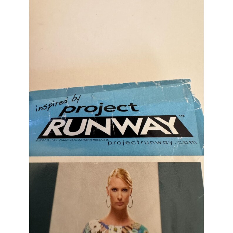 May include: A light blue rectangular paper sign featuring the black and white Project Runway logo with the text inspired by Project Runway and projectrunway.com. Below the logo is a photograph of a person wearing a floral patterned top.