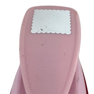 May include: A pink paper punch with a scalloped edge design. The punch has a rectangular silver plate with a scalloped border. The tool is designed for crafting and scrapbooking projects.