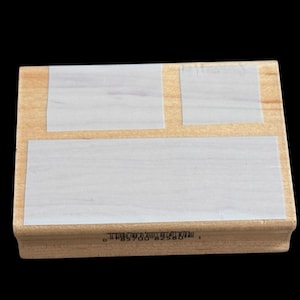 May include: A rectangular wooden rubber stamp block featuring a light brown wood grain finish. The top surface displays three distinct white adhesive labels of varying sizes. A barcode and numerical sequence are printed on the side edge.