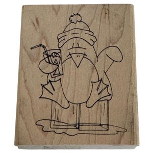 May include: A wooden stamp featuring a cartoon penguin wearing a hat and holding a drink with a straw. The penguin is standing on a block, with a simple outline design in black ink.