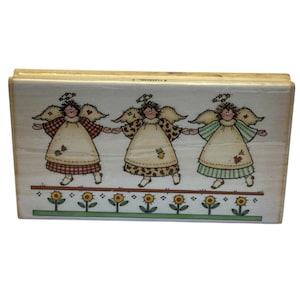 May include: A rectangular wooden rubber stamp featuring an illustration of three angels holding hands. The angels wear dresses with red plaid, brown patterned, and green striped designs. Below them is a row of yellow sunflowers on green stems.