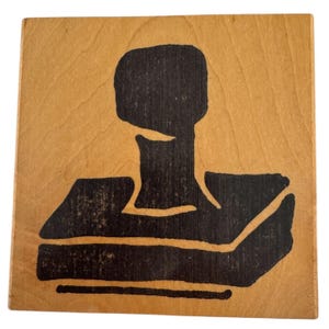 May include: A square wooden block featuring a black ink illustration of a rubber stamp. The natural light brown wood grain is visible throughout the background, while the black graphic depicts the handle and base of a standard office stamp.