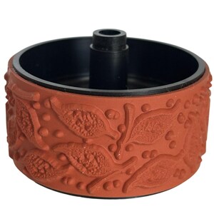 May include: A terracotta-colored stamp with a black interior and a central black post. The stamp's exterior features a repeating pattern of leaves and small circular details. The design is raised, creating a textured surface.
