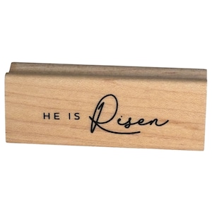 May include: A rectangular wooden rubber stamp featuring a light brown natural wood grain finish. The front surface is laser-engraved with the text HE IS Risen in a combination of bold block lettering and a flowing black cursive script font.