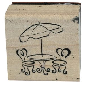 Könnte beinhalten: Ein quadratischer Holzstempel mit einem schwarzen Strichzeichnungsmotiv eines Terrassensets. Das Design zeigt einen runden Tisch mit zwei Stühlen und einem großen Sonnenschirm darüber. Der Stempel besteht aus einem hellen Holzblock.