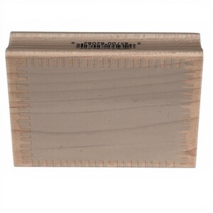 May include: A rectangular wooden rubber stamp block with a light brown wood grain finish. The top edge features a printed barcode and the number 0 85700 82082 0. The center of the block has a smooth, flat surface with a decorative border pattern.