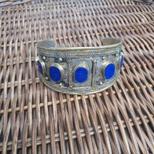 Può includere: Un braccialetto rigido in argento e ottone con pietre blu. Il braccialetto presenta un design decorativo con dettagli intricati.