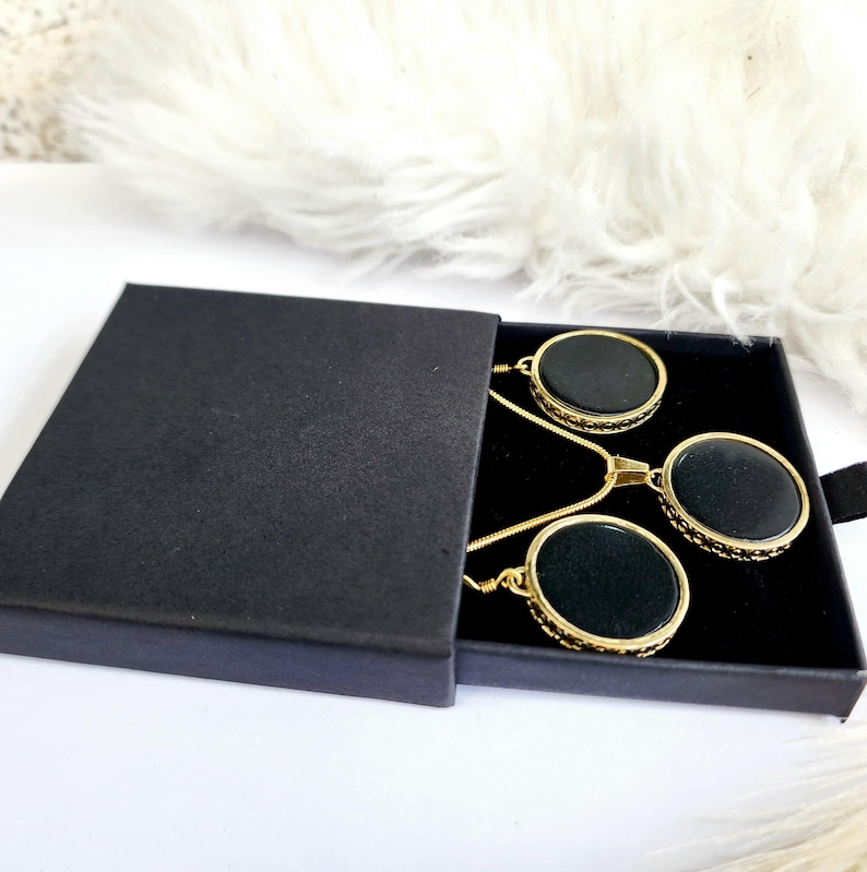 Puede incluir: Un collar de cadena dorada con tres colgantes circulares negros en una caja negra. Los colgantes tienen un borde dorado.