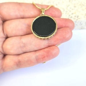 Puede incluir: Un collar de cadena dorada con un colgante redondo negro. El colgante tiene un borde dorado decorativo.