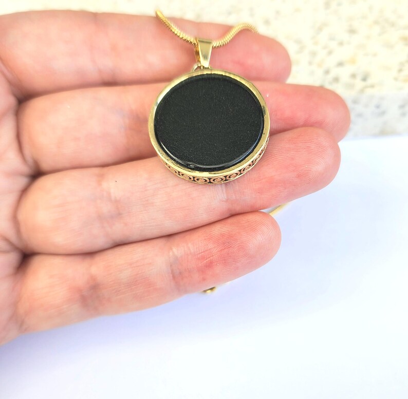 Puede incluir: Un collar de cadena dorada con un colgante redondo negro. El colgante tiene un borde dorado decorativo.