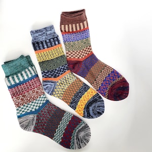 May include: Three pairs of patterned socks in various colors. One pair features a green cuff, red and white geometric patterns, and a gray foot. Another has a blue cuff, orange and yellow sections, and a navy foot. The third pair has a brown cuff and a mix of colors.