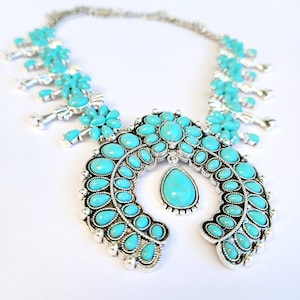 Puede incluir: Un collar turquesa y plateado con un colgante de herradura. El colgante está decorado con piedras turquesa y detalles plateados. El collar es una pieza llamativa y sería perfecto para un estilo western o boho.
