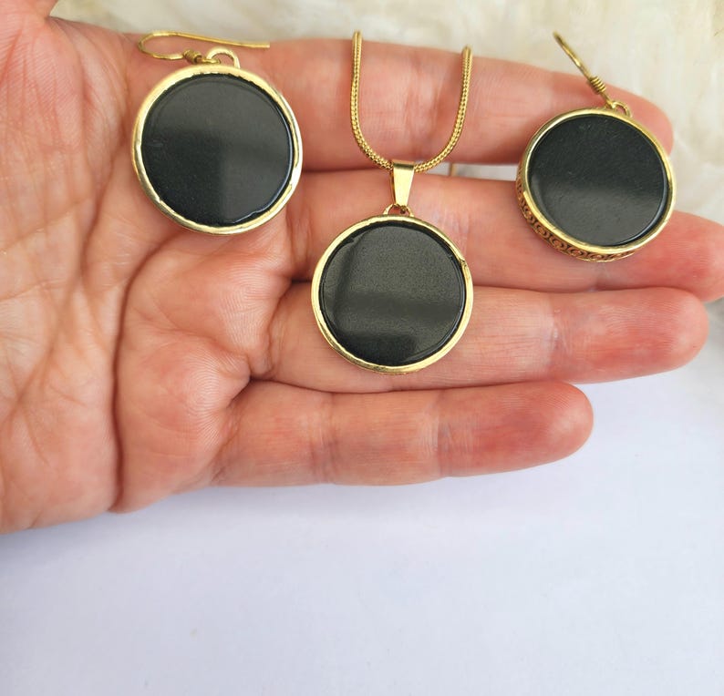 Puede incluir: Un conjunto de collar y pendientes de oro con colgantes redondos negros. Los colgantes tienen una superficie lisa y brillante.