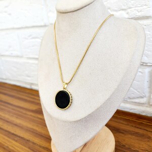 Puede incluir: Un collar de cadena dorada con un colgante circular negro. El colgante tiene un borde dorado con peque&ntilde;os detalles decorativos.