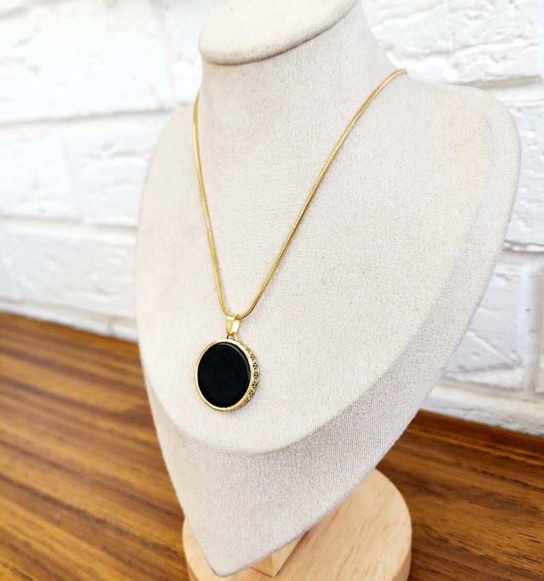 Puede incluir: Un collar de cadena dorada con un colgante circular negro. El colgante tiene un borde dorado con peque&ntilde;os detalles decorativos.