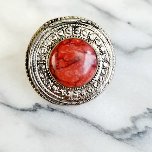 May include: A silver ring with a red and white marbled stone set in the center. The ring has a decorative border with a repeating pattern.
