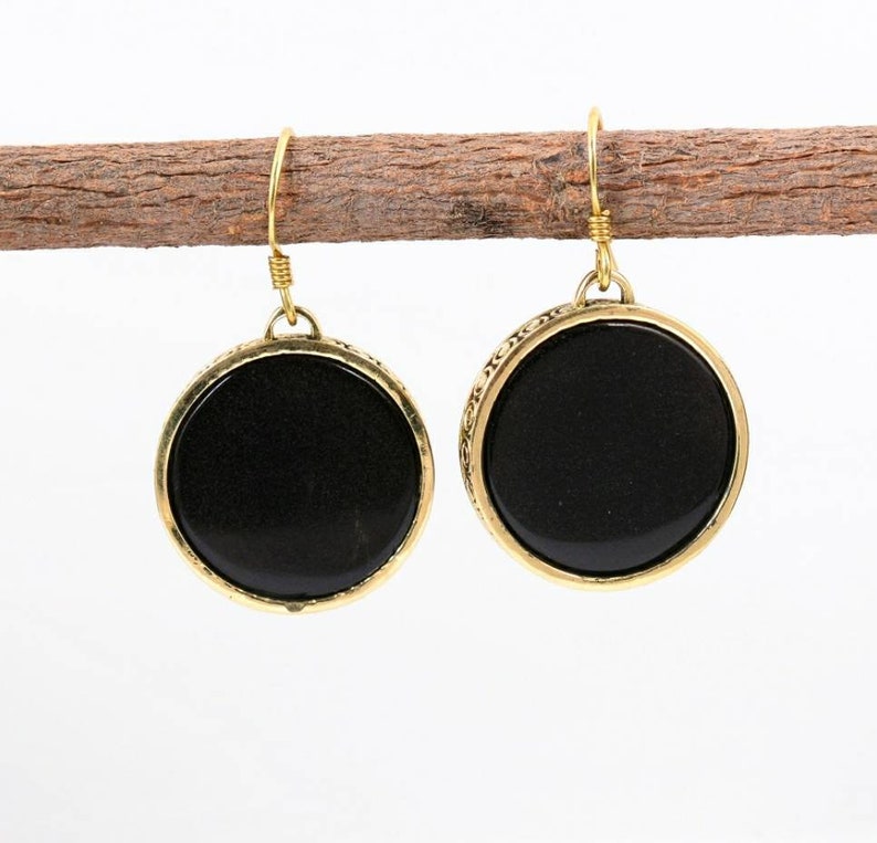 Puede incluir: Un par de pendientes de tono dorado con piedras redondas negras. Los pendientes tienen un dise&ntilde;o decorativo alrededor de las piedras.