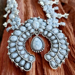Puede incluir: Un collar plateado con un colgante en forma de herradura adornado con piedras veteadas blancas y azules. El collar tiene un diseño floral en la parte superior, sobre un fondo de madera marrón.