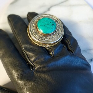May include: A silver ring with a turquoise stone set in a circular design. The ring is on a black leather glove.