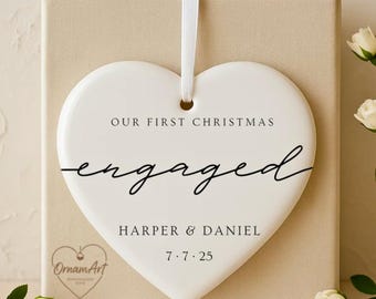 Könnte beinhalten: Ein weißes herzförmiges Keramikornament mit dem Text "OUR FIRST CHRISTMAS engaged HARPER & DANIEL 7.7.25". Das Ornament hängt an einem weißen Band und liegt auf einer beigen Schachtel, mit weißen Rosen.