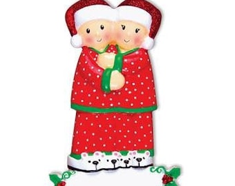May include: A Christmas bauble featuring a couple in red pyjamas with white polka dots, holding hands and wearing matching Santa hats. They are standing on a white base with a blank space for personalisation. The bauble features a holly design and a green ribbon.