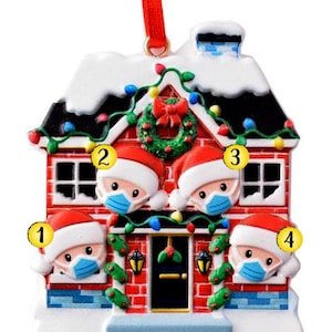 以下が含まれることがあります： サンタの帽子をかぶり、マスクをした4人の人が窓から顔を出している、赤と白の家の形のクリスマスオーナメント。家のドアにはリースが飾られ、屋根には電飾が施されている