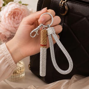 May include: A silver keychain with a lipstick-shaped charm and a wrist strap, both encrusted with rhinestones. The lipstick charm has a gold-colored top and a red band. The wrist strap is a loop of rhinestones. The keychain is held in front of a black purse.