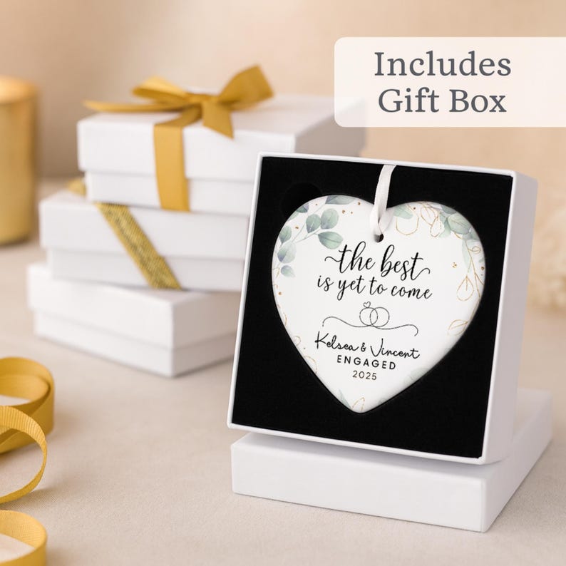 May include: A heart-shaped ceramic ornament with the text "the best is yet to come" and the names "Kelsea & Vincent ENGAGED 2025". The ornament is displayed in a gift box. White gift boxes with gold ribbon are in the background.