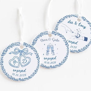 May include: Three white, round engagement tags with blue floral borders and white ribbons. Each tag features unique illustrations and text, including names, dates, and the word "engaged." One tag has two heart-shaped lockets, another has champagne glasses, and the last shows a proposal.