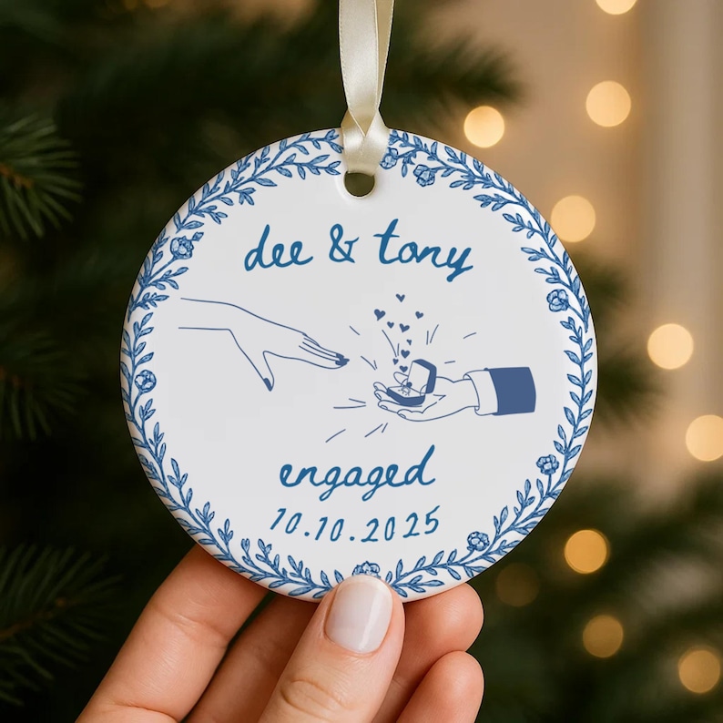 May include: A white ceramic ornament with a blue floral border and the words "dee & tony" above an illustration of a proposal. The word "engaged" and the date "10.10.2025" are also featured. A cream ribbon is attached for hanging.