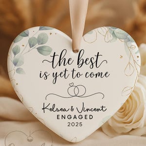 May include: Heart-shaped engagement ornament with the text "the best is yet to come." Features the names Kelsea & Vincent, the word "ENGAGED," and the year 2025. Decorated with floral and gold accents, and a ribbon for hanging.