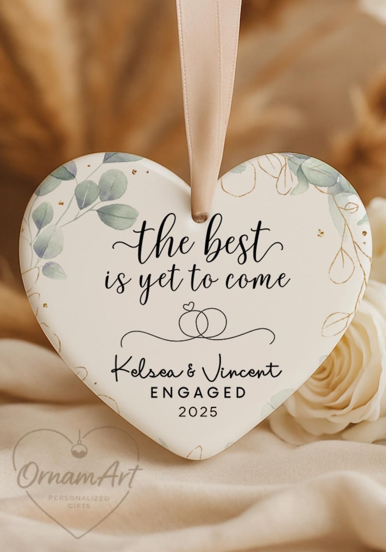 May include: Heart-shaped engagement ornament with the text "the best is yet to come." Features the names Kelsea & Vincent, the word "ENGAGED," and the year 2025. Decorated with floral and gold accents, and a ribbon for hanging.