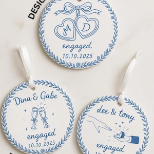 May include: Three white ceramic engagement ornaments with blue designs and white ribbons. Design 1 features intertwined hearts with initials and the date. Design 2 shows champagne flutes. Design 3 depicts a proposal.