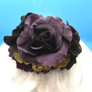 May include: A large, dark purple fabric rose is the focal point of this floral hair accessory. Black, gold, and deep purple fabric petals surround the rose. The accessory is set against a light blue background.